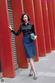 A woman leaning against a red wall wearing a black shirt and blue skirt.